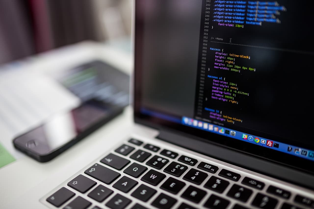 A laptop showing CSS code on screen with a smartphone resting beside it on a desk