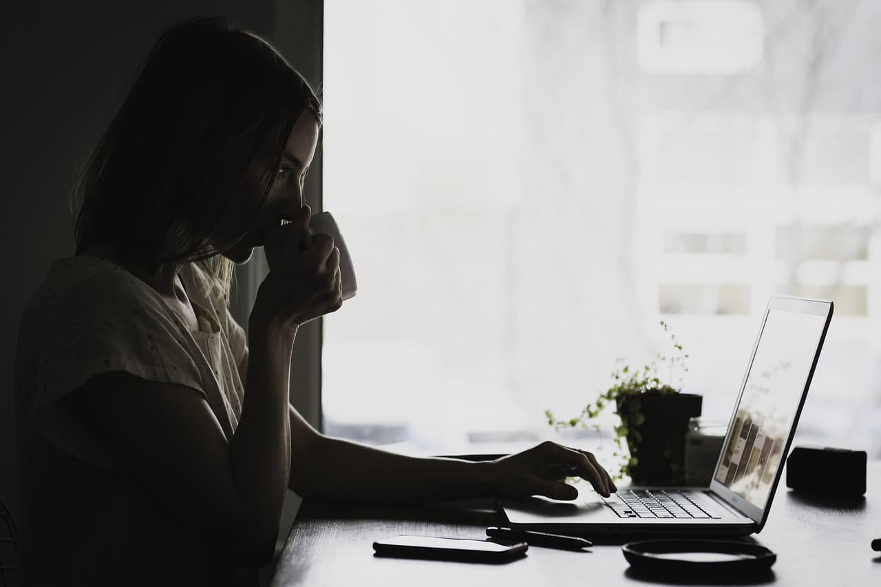 A person at a laptop holding a mug, researching by a window