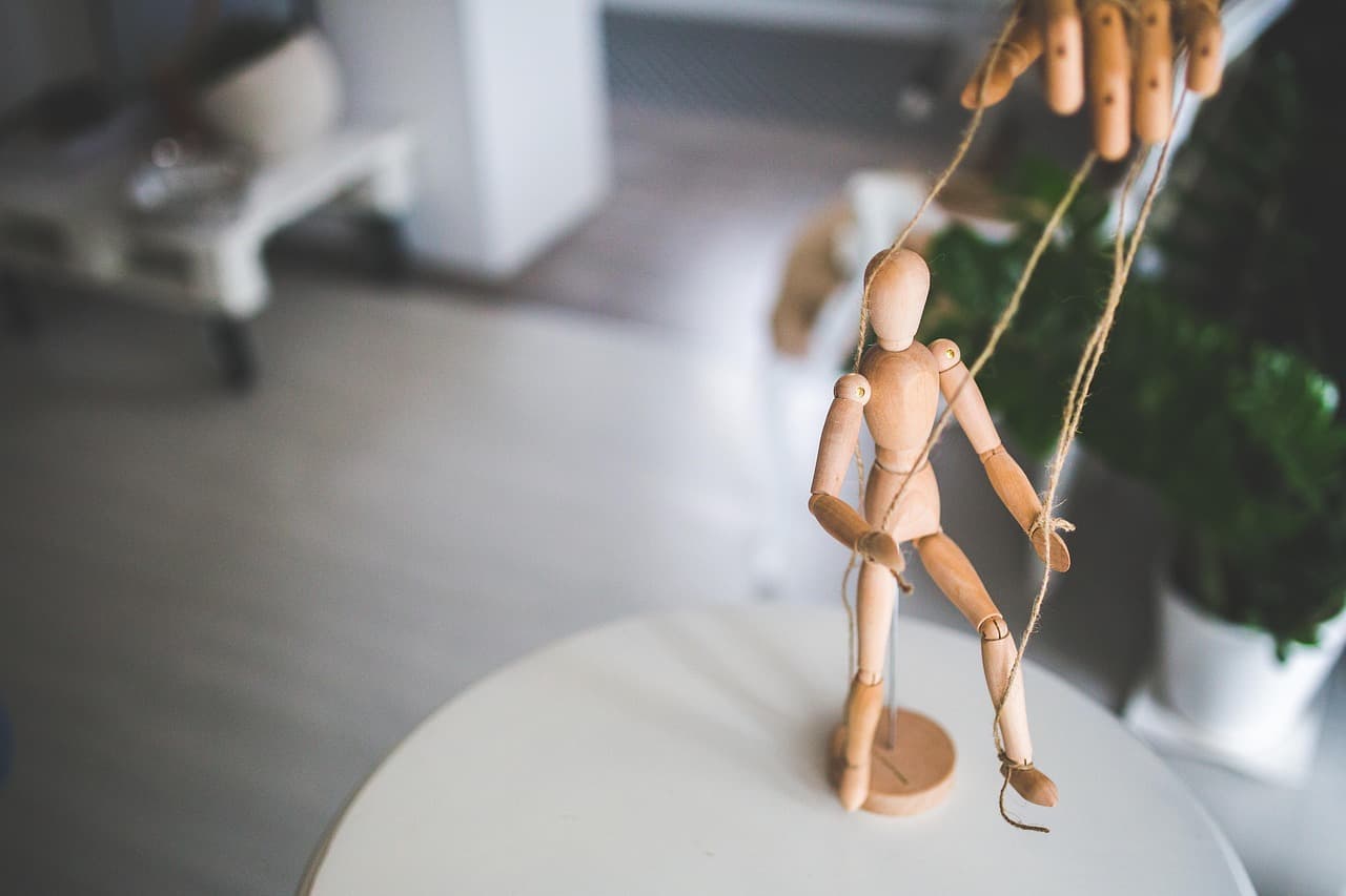 Um manequim de madeira articulado de pé sobre uma mesa branca, com os fios segurados por uma mão humana acima dele