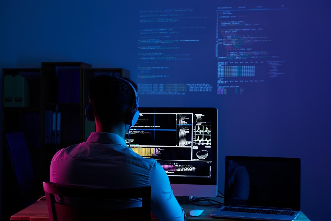 Un développeur au casque travaille à son bureau, du code affiché sur un écran dans une pièce sombre
